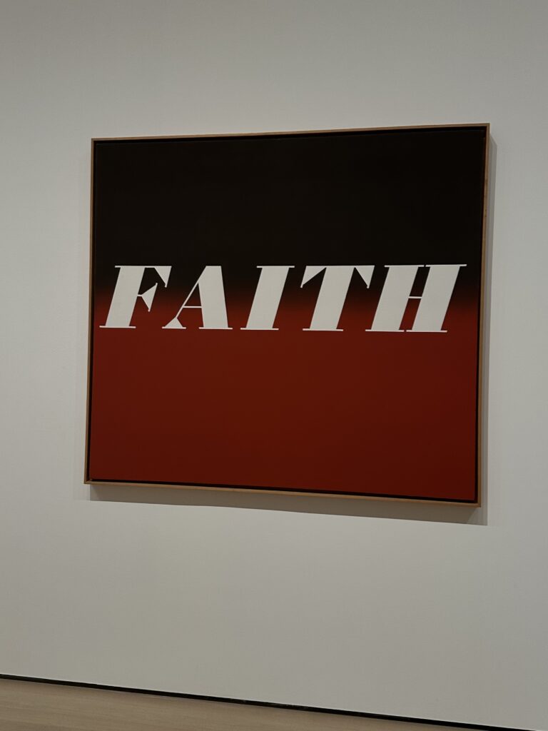 This is a picture at the MoMA of a ombre painting of black to red with “FAITH” written in the middle in white font. 